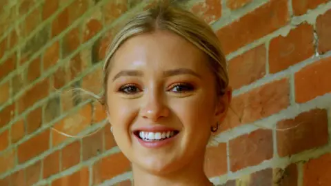 PA Media A young woman with blonde hair tied back. She is smiling at the camera and is stood in front of a brown brick wall