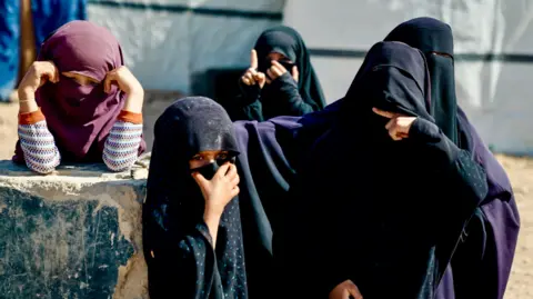 Goktay Koraltan/BBC Veiled women, some are clad head to toe in black. Thee are also a few children. One is raising her index finger. A woman is making a cutting gesture. 