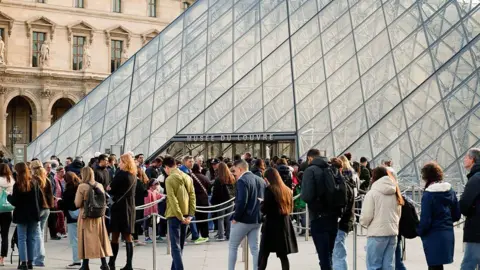 Getty Images A winding queue of visitors outside the entrance to the Louvre, which is a tall glass pyramid intercut with a black criss-crossed pattern.