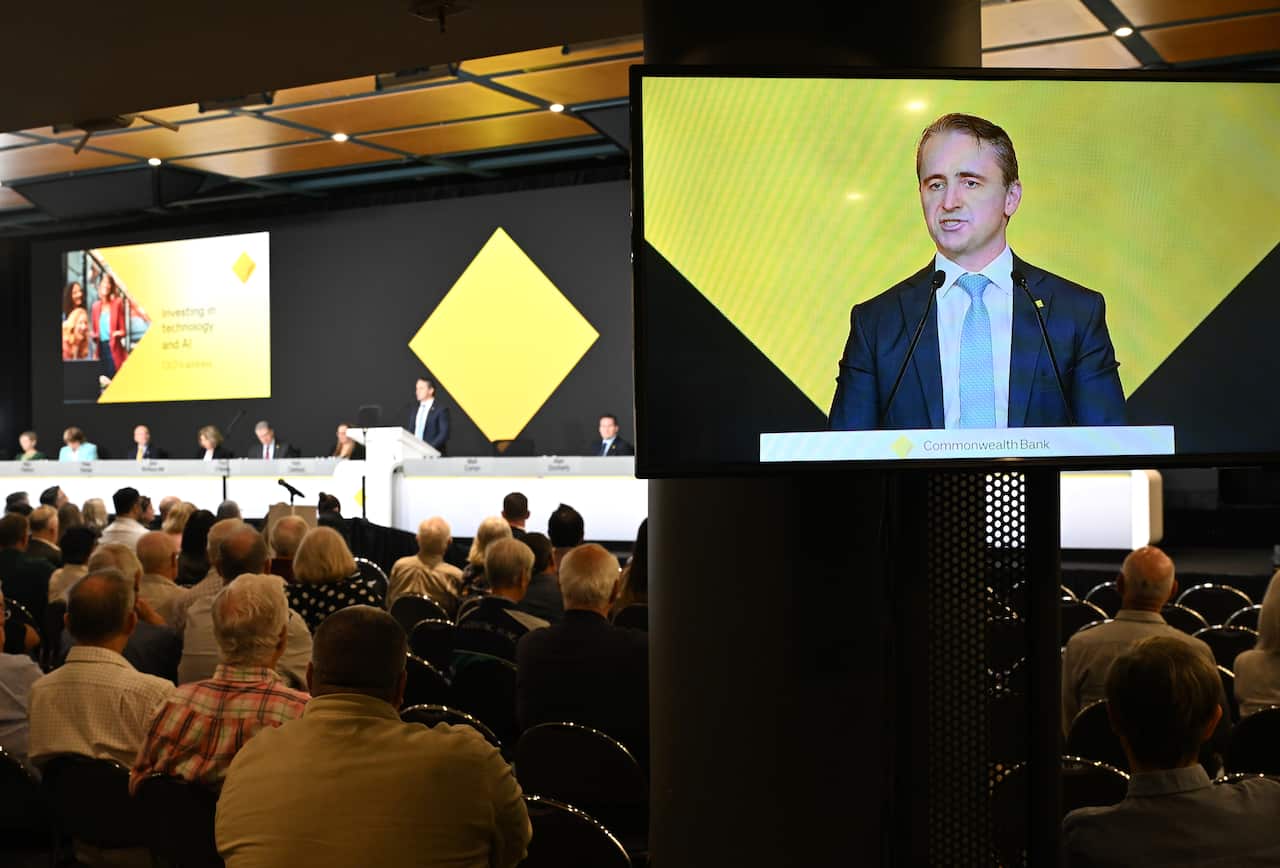 Rows of people are seated, with a panel of people up on stage. A large screen shows a man in a black suit, white shirt and blue tie speaking.