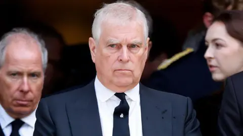 Getty Images Prince Andrew, in dark blue double-breasted suit, white shirt and dark blue tie is standing in front of a man and a woman outside a church. The Prince has white hair and looks very serious.