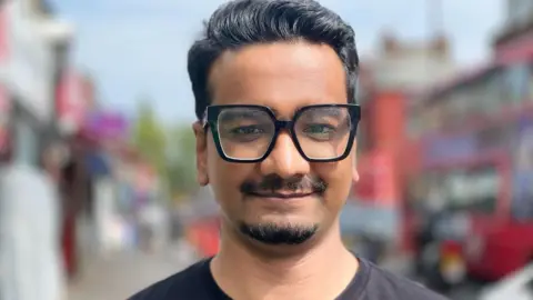 Farhan Jaisin, who is looking at the camera. He has a short goatee beard and moustache with short dark hair and wide-rimmed black glasses. He is wearing a black t-shirt and in the background out of focus there is a London street with a red double decker bus and shops