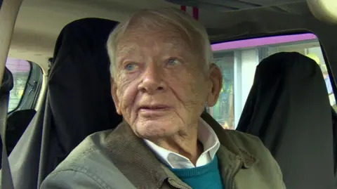 Tommy Foster in a green jacket and blue jumper sitting in a car. He has grey hair. There are black protective covers on the seats.