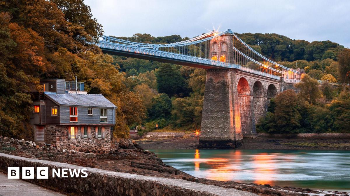 Bridge seen from one side and below, along a path with view of the river below and a house to the left. The bridge is lit in warm yellow light