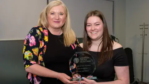 East Ayrshire Council A woman with blond hair and a bright multi-coloured top on, smiling at the camera as she hands an award to a school pupil with long brown hair. Both are looking into the camera and smiling.