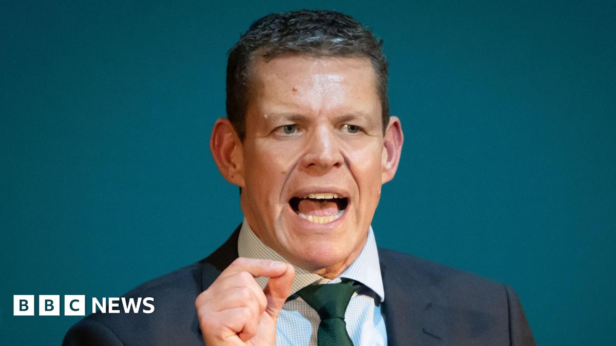 A head and shoulders shot of Rhun ap Iorwerth gesturing with his right hand while he speaks at his conference. He is wearing a black suit and chequered shirt, and a green tie.