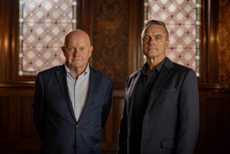 Two middle-aged men in formal attire stand side by side in a historic room with stained glass windows. The man on the left has a bald head and is wearing a navy suit over a light blue shirt. The man on the right has short grey hair and is dressed in a dark suit over a black shirt. The elegant wooden backdrop enhances the sophisticated atmosphere of the setting.