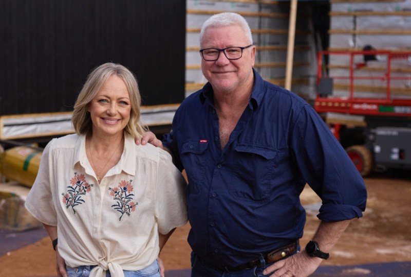 Smiling man and woman posing together at a construction site, with building materials and machinery in the background. The woman is wearing a floral-embroidered shirt and the man is in a navy blue work shirt, showcasing a friendly atmosphere in a renovation or building project.