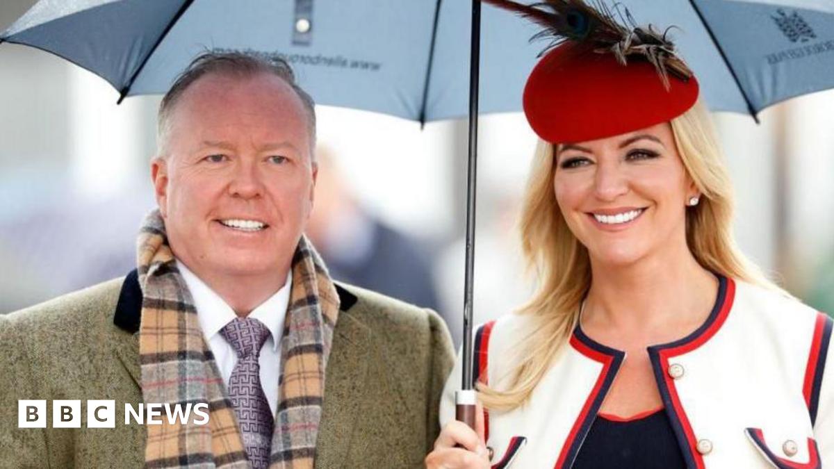 Doug Barrowman and Michelle Mone smartly dressed and smiling sheltering under an umbrella. He wears a tweed coat, checked scarf and purple patterned tie. She wears a red beret with feathers, and a white jacket with black and red trim.