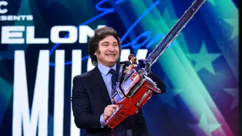 Getty Images Javier Milei wearing a suit wields a chainsaw in front of a big screen while smiling