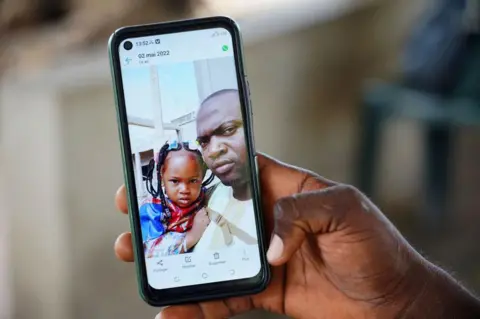 AFP via Getty Images Wuri Bailo Keita, 33, holds a mobile phone showing a picture of himself and his late daughter, Fatoumatta, who is believed to have died of acute kidney failure, in Banjul on October 10, 2022. - Over the last 3 months 69 children have died from acute kidney failure believed to be caused by four cough syrups made by the Indian Pharma company Maiden Pharmaceuticals. Indian authorities are investigating the cough syrups made by the local pharmaceutical company after the World Health Organisation said they could be responsible for the deaths of The Gambian children. (Photo by MILAN BERCKMANS / AFP) (Photo by MILAN BERCKMANS/AFP via Getty Images)