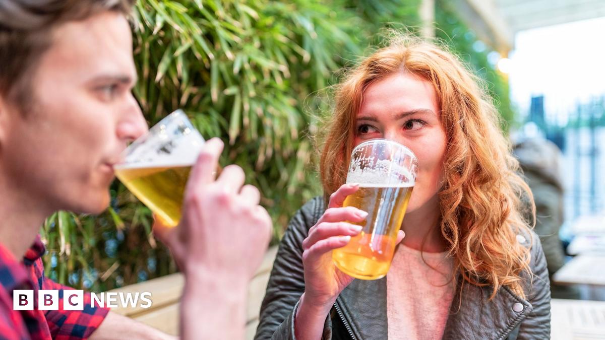 Stock photo shows two people sitting outside drinking pints of beer in front of pub garden foliage, one has short brown hair, the other light ginger hair.