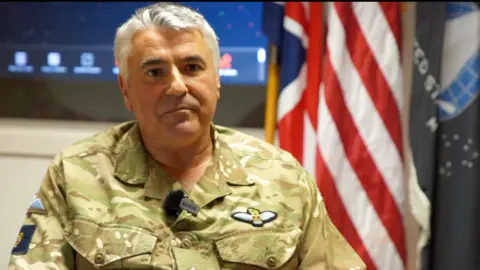 The head of the UK Space Command sits looking at the camera, wearing a camouflage military uniform with flags in the background.