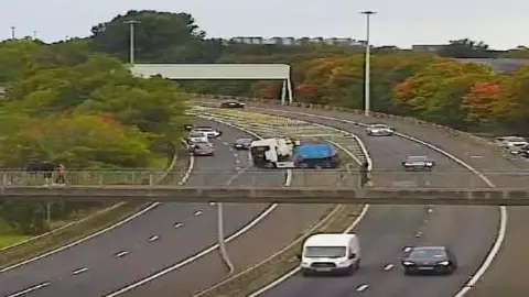 Traffic Scotland A lorry cab in the middle verge between two double lanes of traffic with a footbridge in the foregound, autumn trees behind and several vehicles driving by