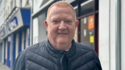 Bill Pedan in a puffer jacket and jumper standing on the street. He has short red hair. There are shops behind him.