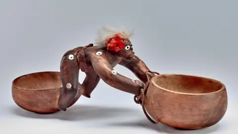 Trustees of the British Museum Two carved wooden bowls linked by a carved wooden human figure.