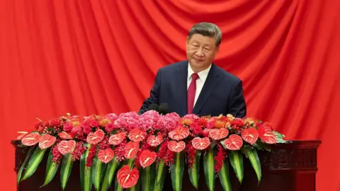 Reuters Xi Jinping wearing a dark suit stands in front of a red curtain and behind a wooden lectern covered in bright red and pink flowers and green leaves