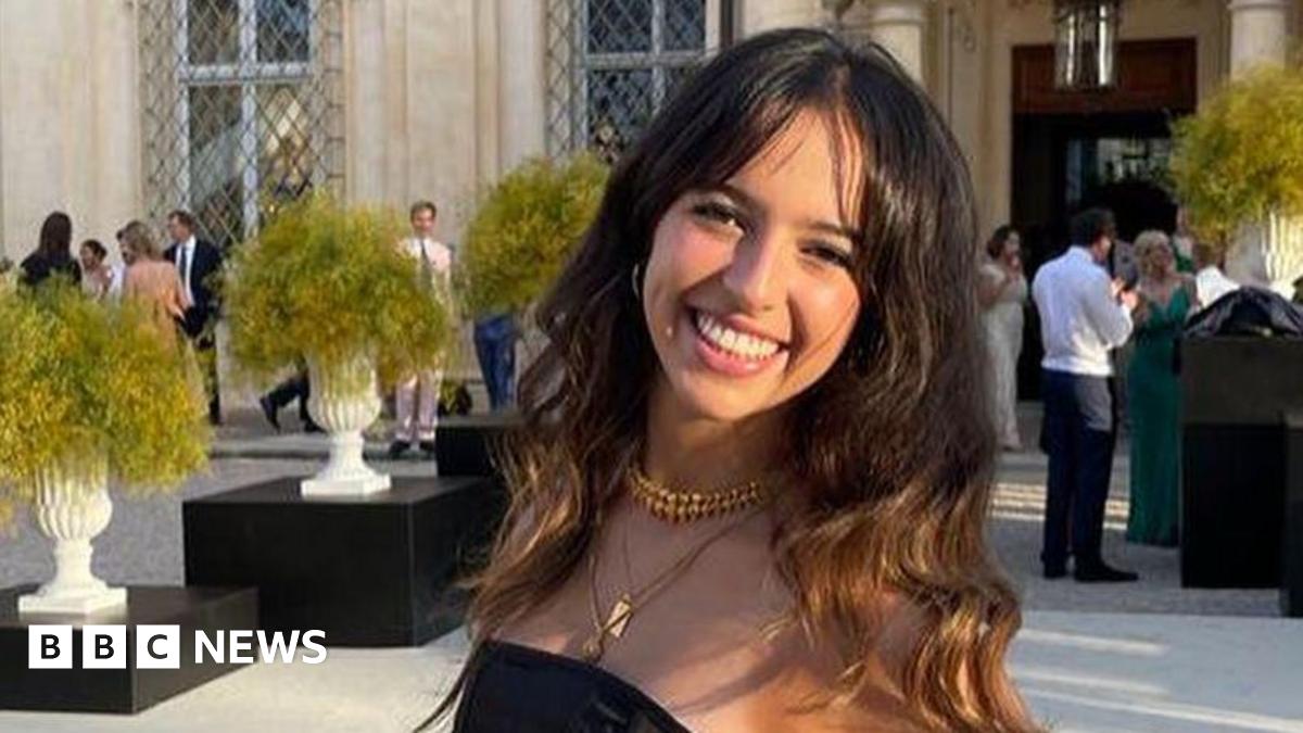 Kemi Badenoch wearing a white jacket and white t-shirt is pictured waving to the camera. There is a union jack pictured to the left.