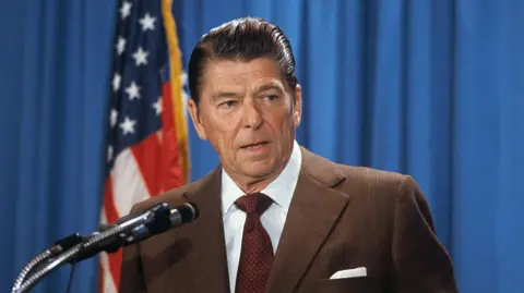 Getty Images A file photo of Ronald Reagan from the 1970s. He wears a brown suit jacket, a dark red tie, a white shirt, and has slicked back brown hair. He speaks into a microphone and stands in front of a US flag and a blue curtain.