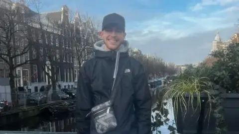 Hertfordshire Constabulary Robert Tyler-Jones standing on a bridge, in front of a canal and buildings, smiling at the camera, wearing a black cap, black top, trousers and a bag across him. He is standing next to a plant display.