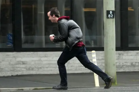 Getty Images A man wearing a black coat and jeans, carrying a coffee cup, struggling to cross a road during a strong wind