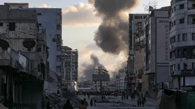 Smoke rises over Gaza City, as a crowd of Palestinians stands below in the street