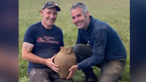 Minelab Two men holding a clay pot smiling with the dig site seen just in front of them. The man on the left has glasses on his head and a cap, he has a blue t-shirt with the words Ben Sherman, in red font, written on the front. The man on the right has salt and pepper hair, a blue shirt and blue pants. 