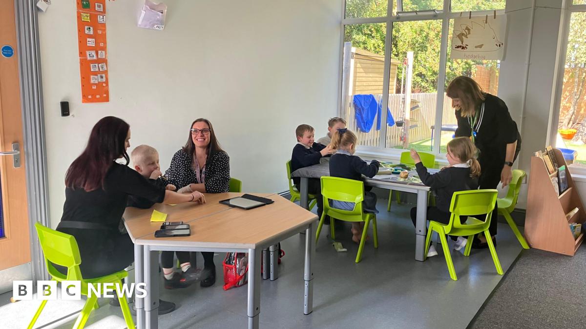 Five pupils aged below six, plus three staff sit across two tables with green chairs in a classroom setting. One table is working with technology, and the other table is chopping vegetables.