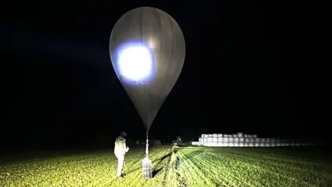 State Border Guard Service via AP A weather balloon that has landed in a field, at night, is inspected by an official 