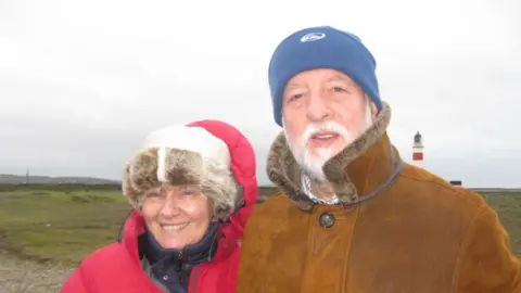 SUSAN JOHNSON Muriel, a woman with fluffy hat, red coat and a bright smile, is photographed standing next to her husband Barrie, a man with a white beard and moustache, a blue hat and brown coat.