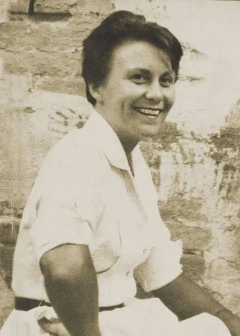 Michael Brown Sepia photo of a smiling Harper Lee looking to camera -- side on. with her right arm on her waist. She's wearing a white short sleeved blouse and white skirt.
Background og brick wall.