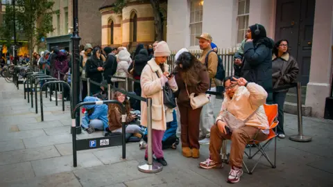 A line of a dozen or so people with one sitting on a chair showing a peace sign, others looking at phones and some sitting on the ground