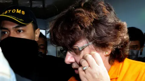 Reuters Lindsay Sandiford of Britain is escorted by custom officers before a news conference at the Customs Office at Ngurah Rai International Airport in Bali. She has short brown hair, glasses and a yellow shirt. She is holding her hand to her face.