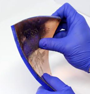 Closeup of a pair of gloved hands bending a chip-sized processor and 3D printed antenna array.