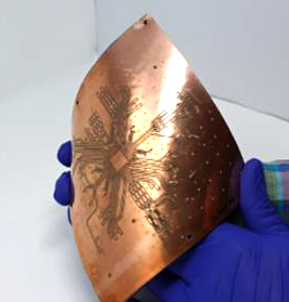 Closeup of a pair of gloved hands bending a chip-sized processor and 3D printed antenna array.