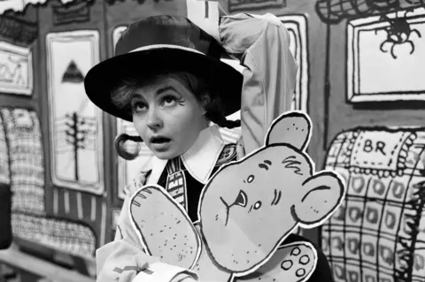 Getty Images Prunella Scales photographed in one of her earliest acting roles in 1962. She is dressed as a school girl with pigtails and a boater hat.  She is carrying a cut-out of a teddy bear.