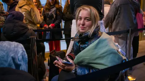 Sophie sits on a chair with a blanket around her and phone in her hand