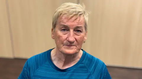 BBC A woman with short blonde hair is looking into the camera. She is wearing a blue striped top and gold hoop earrings.  