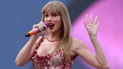 PA Media Taylor Swift with long blonde hair and a fringe. She is performing on stage with a pink and orange microphone in hand and she is wearing a jewelled costume.