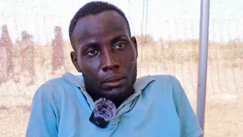 BBC A head and shoulders shot of man in a light blue shirt. He has a clip mic attached to a collar. 