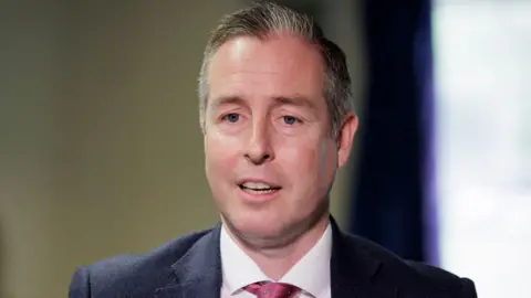 Liam McBurney/PA Wire Paul Givan, a man with blue eyes and short grey hair, looks towards the camera.  He is mid-speech and is wearing a navy suit, a white shirt and a pink tie. 
