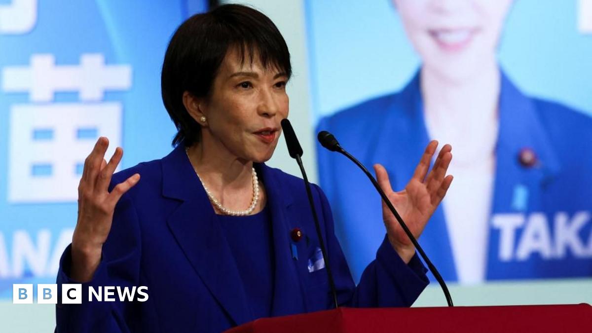 Sanae Takaichi gestures with both hands as she speaks into a pair of microphones at a red lectern. She wears a royal blue suit, pearl earrings and a pearl necklace. She stands in front of a screen on which is projected a picture of her smiling, again wearing blue, alongside her surname, printed in large, white capital letters.