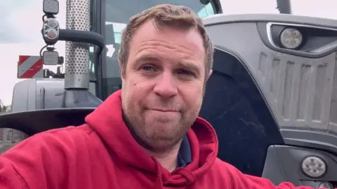 Olly Harrison Olly Harrison has short hair and a short beard. He is wearing a red hooded top and is stood in front of a grey tractor.