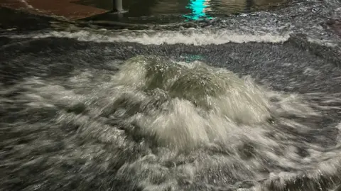 Dan Jessup Water surges out of a manhole cover on a road. 