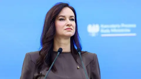 Getty Images Inga Ruginiene, a woman with waved dark hair, stands at a podium with two small microphones in front of a blue background, wearing a chocolate brown long-sleeved dress