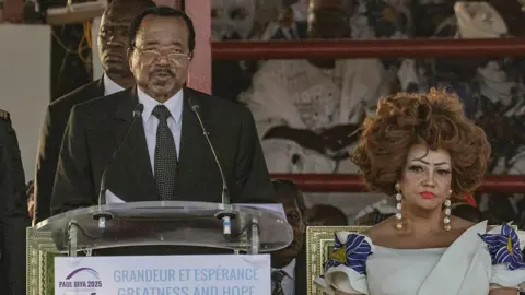 AFP President Paul Biya stands at a lectern and speaks into a pair of microphones. He is flanked by his wife, Chantal Biya. 