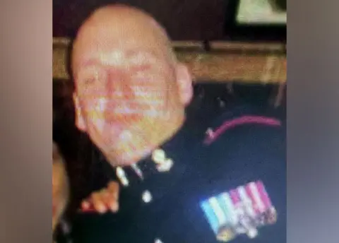 Facebook A blurred image of Michael Webber - he's wearing a dark-coloured uniform and has medals on the left side with ribbons of purple/white and blue/white. He's bald.
