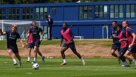 Wycombe Wanderers The team trains on an outdoor pitch with a blue building in the background. About 9 players wearing pink bibs over blue kit are chasing the ball.