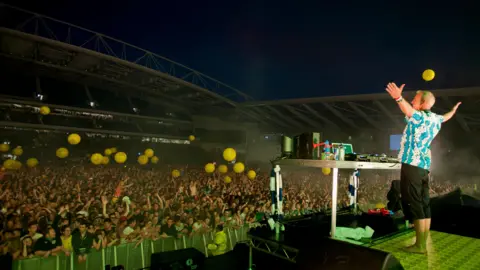 Getty Images A man in three-quarter length trousers and a blue and white shirt stands behind a table with DJ decks, arms outstretched, facing a large crowd.