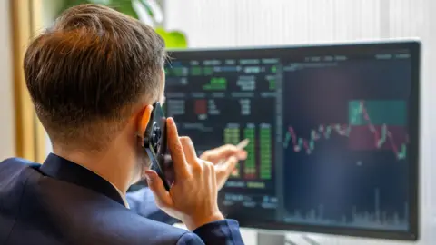 Getty Images A man with his back to the camera holds a mobile phone to his ear and points to computer screens showing financial market information
